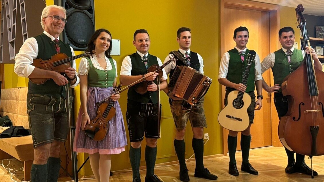 Die in traditioneller Tracht gekleidete Gruppe der Ausseer Bradlmusi in der großen Besetzung von 6 Musikanten und Musikantinnen hält ihre Instrumente in der Hand, sie stehen herzlich lächelnd gemeinsam in einem Raum mit schönem Parkettboden, Spotlights und großem Lautsprecher im Hintergrund. | © Ausseer Bradlmusi