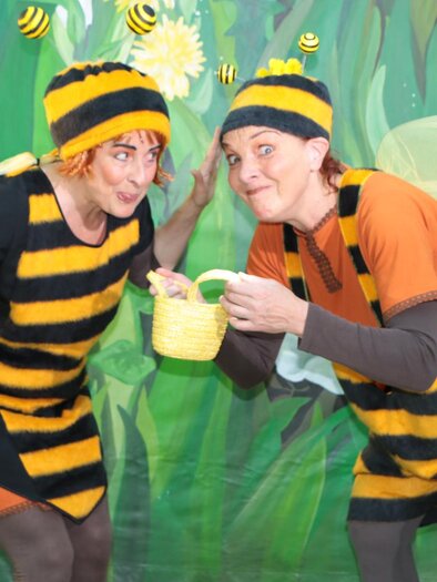 Zwei Personen in bunten Bienenkostümen stehen vor einer grünen Blumenwand. Sie haben fröhliche Gesichtsausdrücke und scheinen miteinander zu kommunizieren. | © ©KK