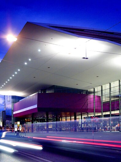 Ein modernes Gebäude bei Dämmerung, das mit einer großen Glasfassade und einem auffälligen Dachdesign beeindruckt. Die Lichter der Umgebung reflektieren sich auf der Oberfläche und erzeugen eine dynamische Atmosphäre. | © mcg_werner krug