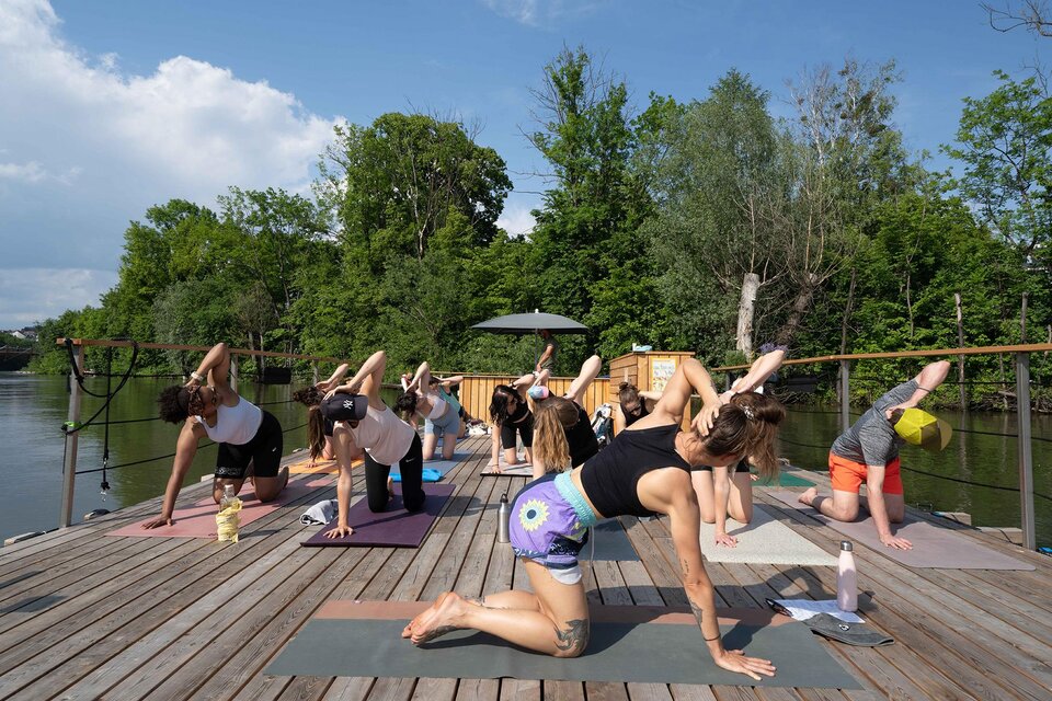 Eine Gruppe von Menschen macht Yoga auf einem Holzdeck am Wasser. Umgeben von Grün und unter einem blauem Himmel praktizieren sie verschiedene Yoga-Posen. | © Flösserei