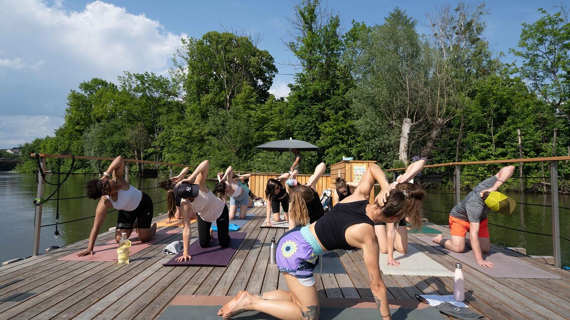 Eine Gruppe von Menschen macht Yoga auf einem Holzdeck am Wasser. Umgeben von Grün und unter einem blauem Himmel praktizieren sie verschiedene Yoga-Posen. | © Flösserei