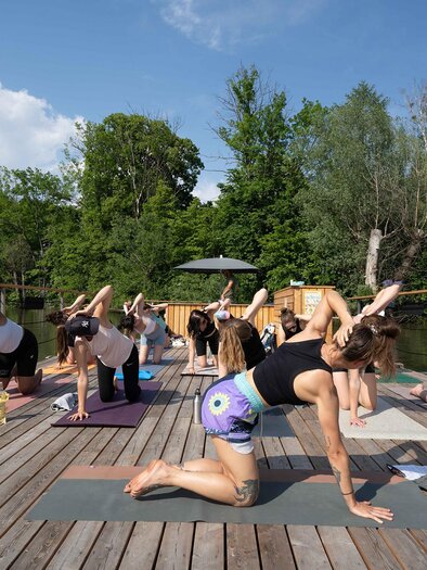 Eine Gruppe von Menschen macht Yoga auf einem Holzdeck am Wasser. Umgeben von Grün und unter einem blauem Himmel praktizieren sie verschiedene Yoga-Posen. | © Flösserei