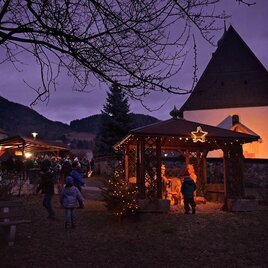 Ein festlicher Weihnachtsmarkt mit beleuchteten Ständen und einer Holzhütte in der Dämmerung. Menschen schlendern gemütlich vorbei, während die Umgebung festlich dekoriert ist.