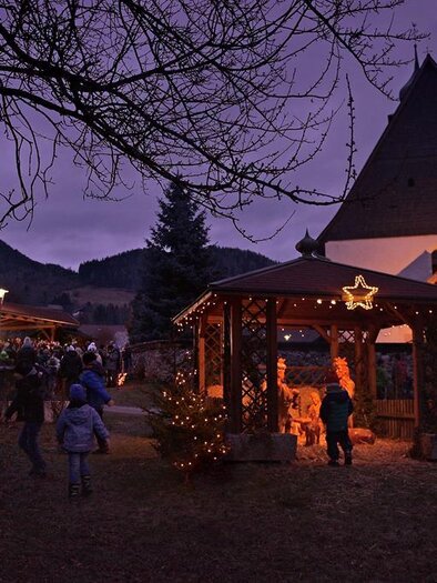 Ein festlicher Weihnachtsmarkt mit beleuchteten Ständen und einer Holzhütte in der Dämmerung. Menschen schlendern gemütlich vorbei, während die Umgebung festlich dekoriert ist.