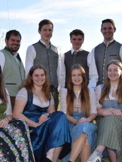 Eine Gruppe von Personen in traditioneller Tracht steht und sitzt zusammen im Freien. Der Hintergrund zeigt einen sonnigen Himmel und eine grüne Landschaft. | © Landjugend St. Peter
