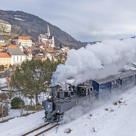 Eine Dampflok fährt durch eine verschneite Landschaft mit einem kleinen Dorf im Hintergrund. Die Szene zeigt eine malerische Winterlandschaft mit Schnee und Dampf. | © Tv Murau