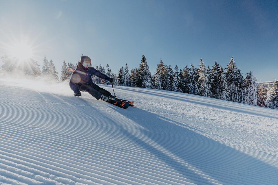 Ein Skifahrer fährt mit Geschwindigkeit eine schneebedeckte Piste hinunter. Im Hintergrund sind schneebedeckte Bäume und ein klarer blauer Himmel zu sehen. | © nicoleseiser.at