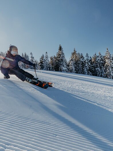 Ein Skifahrer fährt mit Geschwindigkeit eine schneebedeckte Piste hinunter. Im Hintergrund sind schneebedeckte Bäume und ein klarer blauer Himmel zu sehen. | © nicoleseiser.at