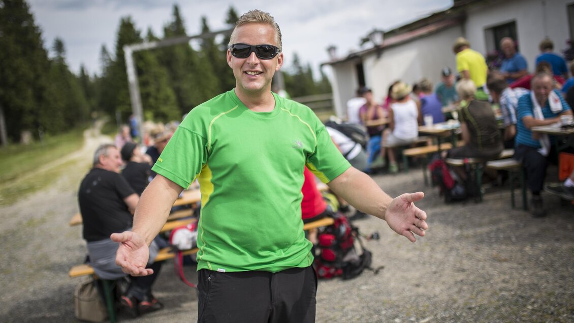 Ein Mann in einem grünen Sportshirt lächelt und öffnet die Arme. Im Hintergrund sind viele Personen an Tischen versammelt. | © Die Edlseer/BePo