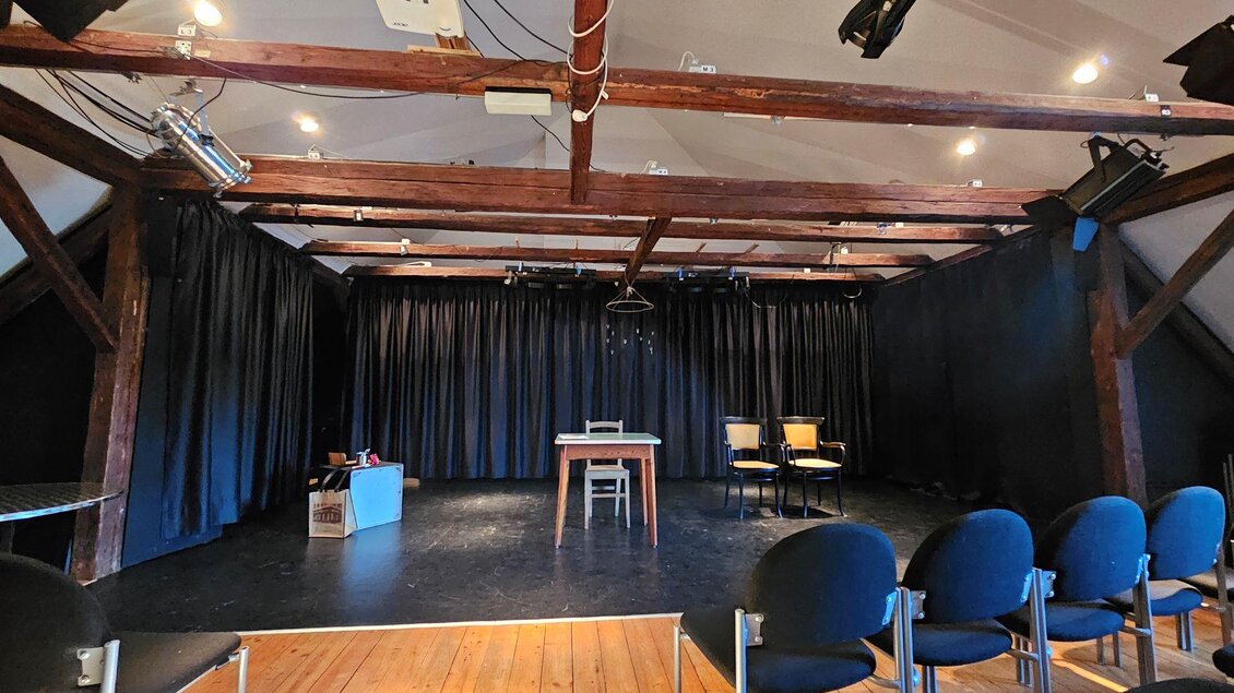 Ein kleines Theater mit dunklen Vorhängen und einer leeren Bühne. Zwei Stühle und ein Tisch stehen auf der Bühne, während die Sitzreihen aus schwarzen Stühlen bereit sind. | © StainZeit - Peter Nöhrer