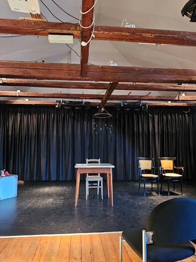 Ein kleines Theater mit dunklen Vorhängen und einer leeren Bühne. Zwei Stühle und ein Tisch stehen auf der Bühne, während die Sitzreihen aus schwarzen Stühlen bereit sind. | © StainZeit - Peter Nöhrer