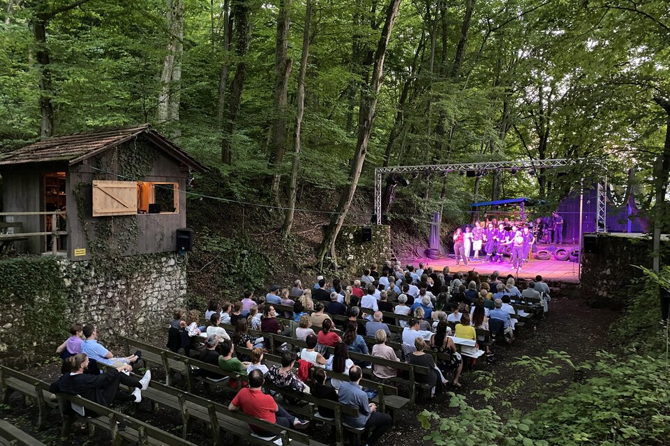 Eine Open-Air-Veranstaltung im Wald mit einer Bühne und Publikum. Umgeben von Bäumen, sitzen die Zuschauer auf Holzbänken. | © Wildoner Schlossbergbühne