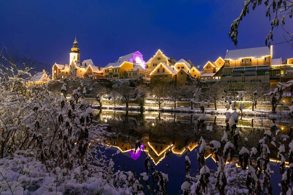 Eine malerische Winterlandschaft mit verschneiten Bäumen und einem ruhigen Fluss. Im Hintergrund erstrahlen die Häuser in festlicher Beleuchtung. | © Region Graz-Harry Schiffer