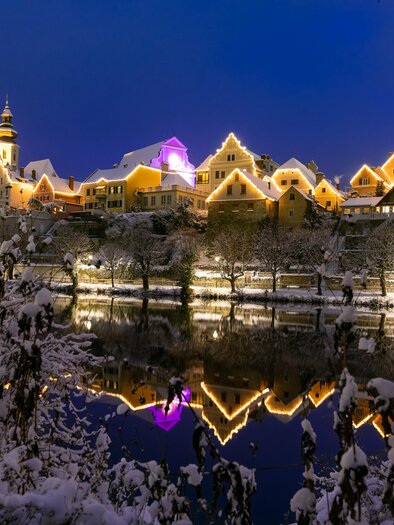 Eine malerische Winterlandschaft mit verschneiten Bäumen und einem ruhigen Fluss. Im Hintergrund erstrahlen die Häuser in festlicher Beleuchtung. | © Region Graz-Harry Schiffer