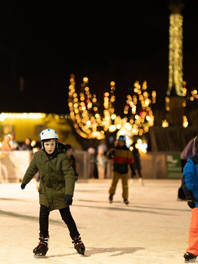 Eine Eislaufbahn bei Nacht mit vielen fröhlichen Menschen. Kinder skaten in bunten Jacken unter funkelnden Lichtern.