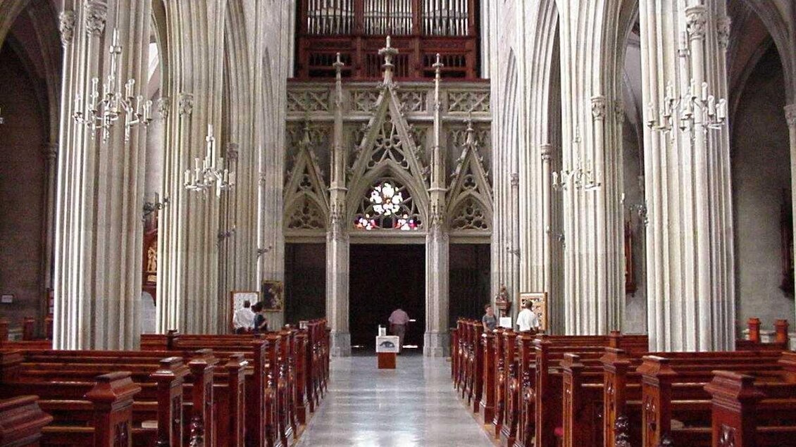 Eine beeindruckende Kircheninneneinrichtung mit hohen gewölbten Decken und eleganten Holzbänken. Im Hintergrund befindet sich eine große Orgel und ein schönes Kirchenfenster.