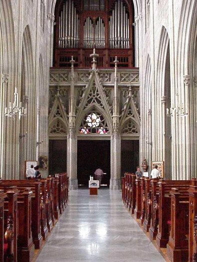 Eine beeindruckende Kircheninneneinrichtung mit hohen gewölbten Decken und eleganten Holzbänken. Im Hintergrund befindet sich eine große Orgel und ein schönes Kirchenfenster.