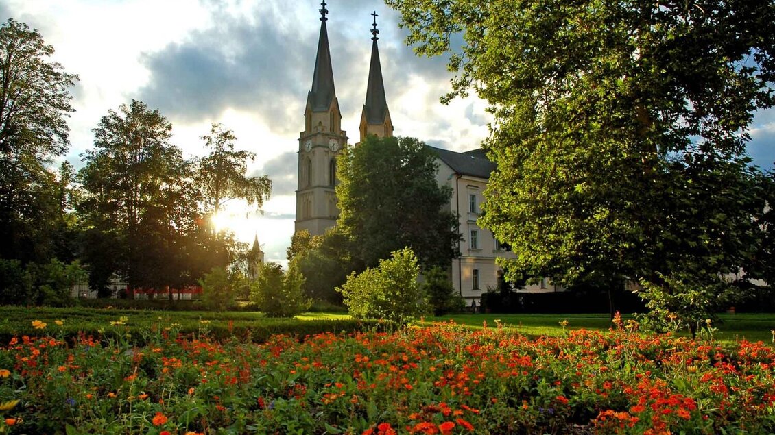 Ein schöner Garten mit bunten Blumen und grünen Bäumen. Im Hintergrund sind hohe Kirchtürme und ein bewölkter Himmel zu sehen. | © Thomas Sattler