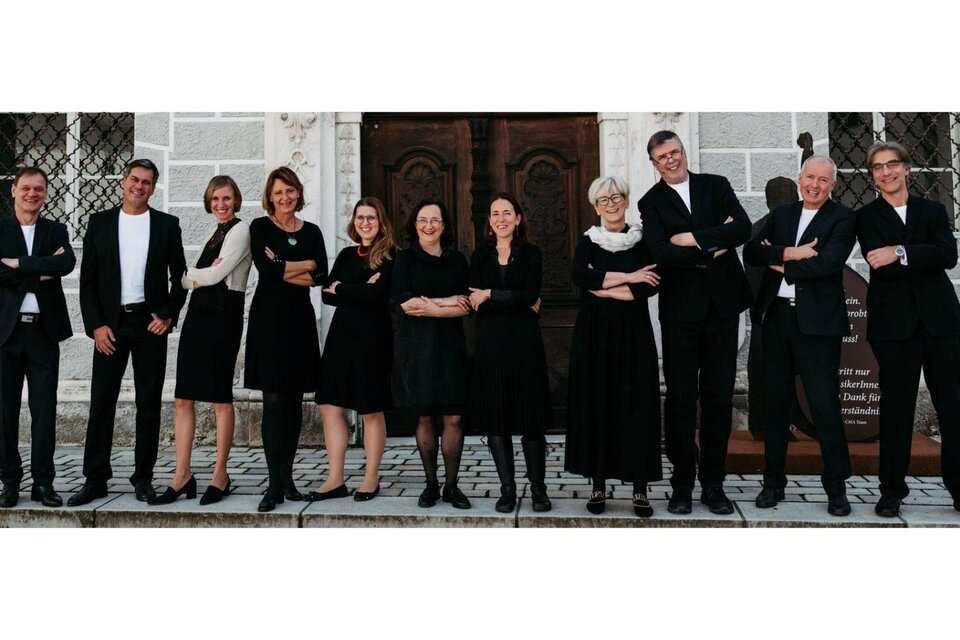 Eine Gruppe von zehn Personen steht vor einem historischen Gebäude. Alle tragen schwarze Kleidung und lächeln, während sie sich in einer lockeren Pose präsentieren. | © Ensemble Vokal