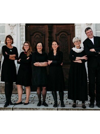 Eine Gruppe von zehn Personen steht vor einem historischen Gebäude. Alle tragen schwarze Kleidung und lächeln, während sie sich in einer lockeren Pose präsentieren. | © Ensemble Vokal