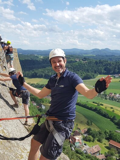 Ein Mann klettert an einer Felswand und lächelt in die Kamera. Im Hintergrund sind grüne Felder und ein blauer Himmel zu sehen. | © Naturbursch Training Riegersburg