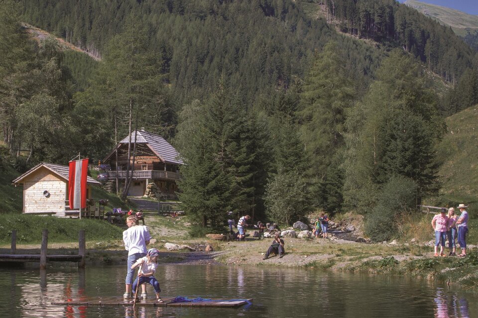 Ein malerischer See mit Familien, die Freizeitaktivitäten genießen. Im Hintergrund sind Bäume und traditionelle Hütten zu sehen. | © Goldwaschanlage Pusterwald
