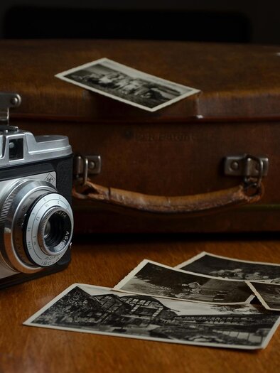 An old camera lies next to a brown travel bag on a table. Several black-and-white photos are scattered around it. | © Pixabay