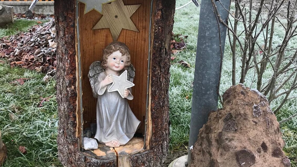 Eine hölzerne Skulptur mit einem Engel, der ein Buch hält, steht in einer rustikalen Umgebung. Daneben sind Steine und eine Metallstütze zu sehen. | © Marktgemeinde Halbenrain