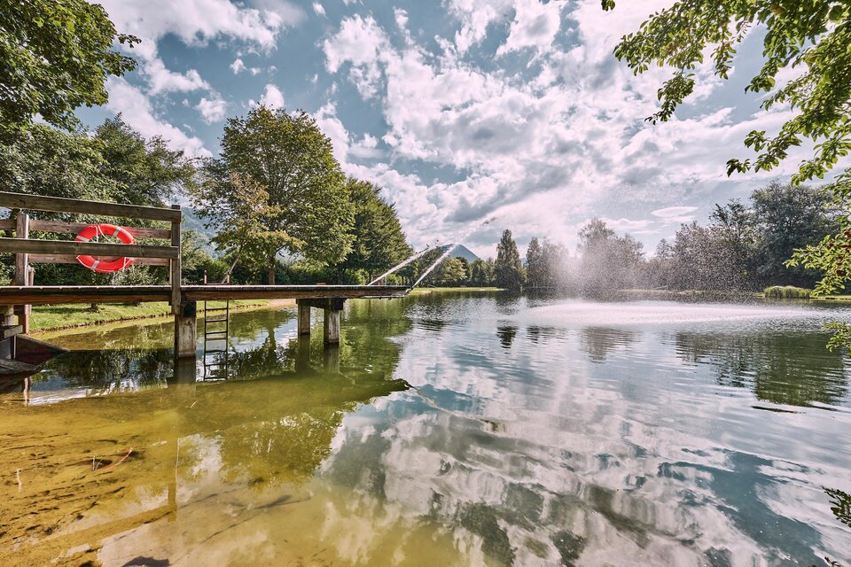 Ein ruhiger See mit einem Steg und einem Schwimmring. Die Wolken spiegeln sich im Wasser, umgeben von grünen Bäumen. | © Thomas Sattler