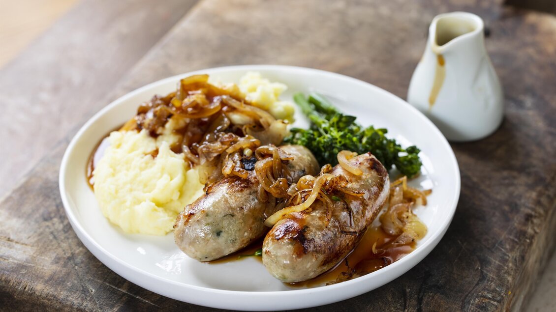 Ein Teller mit Bratwürsten, Zwiebeln, Kartoffelpüree und Brokkoli. Dazu gibt es eine kleine Sauce in einer weißen Kanne. | © Adobe Stock