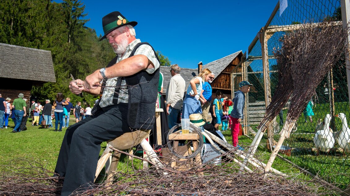 Ein älterer Mann sitzt im Freien und fertigt kunsthandwerkliche Gegenstände aus Zweigen. Im Hintergrund sind Menschen und traditionelle Gebäude zu sehen. | © Region Graz-Harry Schiffer