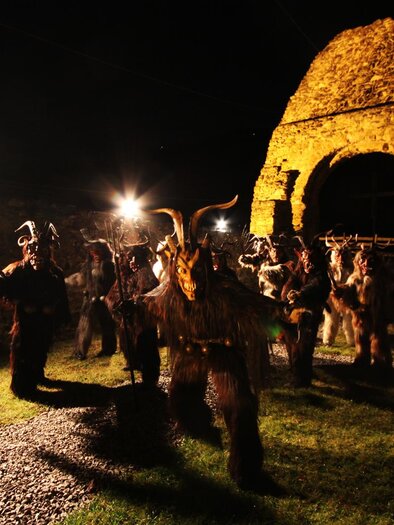 Eine Gruppe von Personen in traditionellen Kostümen steht in einem dunklen Bereich vor einer alten Ruine. Helle Scheinwerfer beleuchten die Szene und schaffen eine festliche Atmosphäre. | © Brauchtumsgruppe Erlsberg