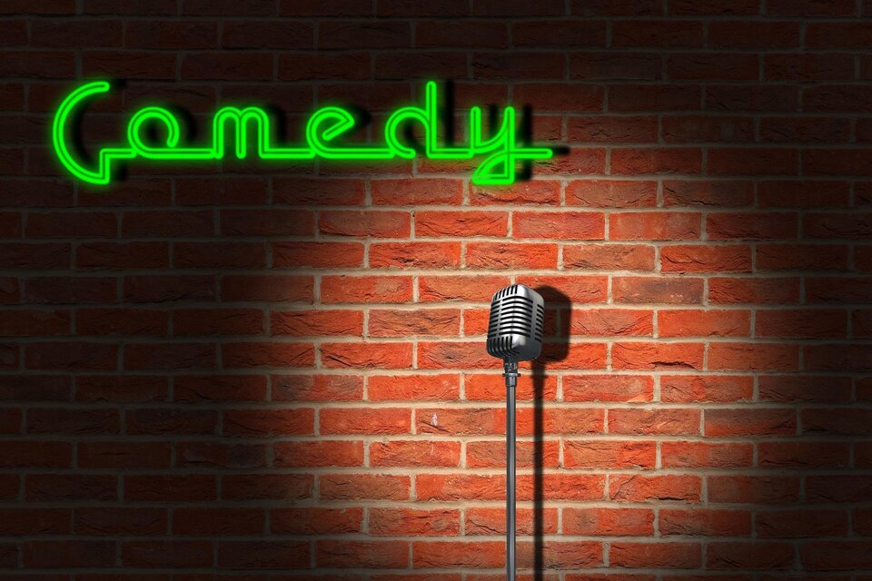 Ein Mikrofon steht vor einer Ziegelwand mit dem grünen Neonlicht, das das Wort „Comedy“ zeigt. Die Szene vermittelt eine einladende Atmosphäre für Comedy-Auftritte.