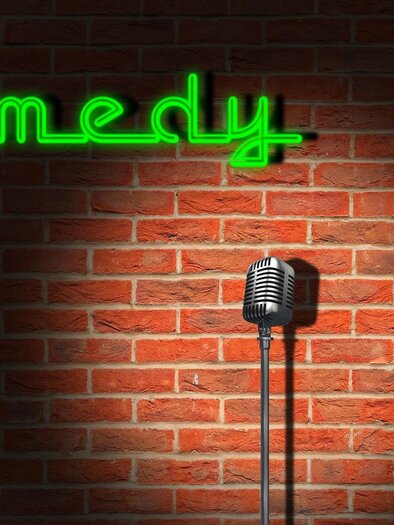 Ein Mikrofon steht vor einer Ziegelwand mit dem grünen Neonlicht, das das Wort „Comedy“ zeigt. Die Szene vermittelt eine einladende Atmosphäre für Comedy-Auftritte.