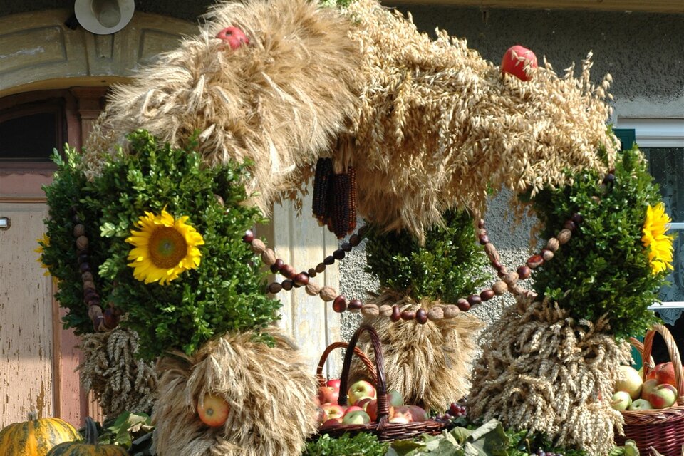 Ein festlich dekorierter Erntekranz aus Heu, Pflanzen und Sonnenblumen. Im Hintergrund ist ein Gebäude mit einem roten Dach zu sehen. | © TV Südsteiermark