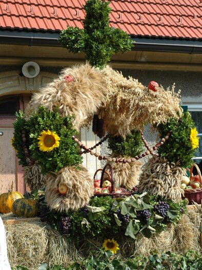 Ein festlich dekorierter Erntekranz aus Heu, Pflanzen und Sonnenblumen. Im Hintergrund ist ein Gebäude mit einem roten Dach zu sehen. | © TV Südsteiermark