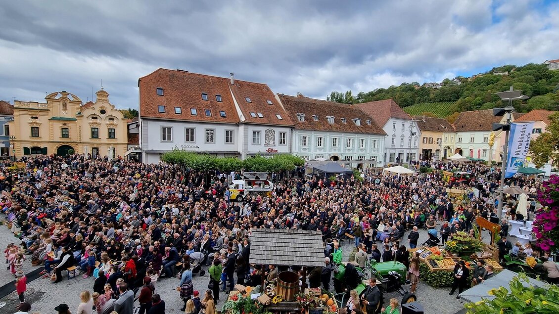 Eine große Menschenmenge versammelt sich auf einem Platz mit bunten Ständen. Über der Veranstaltung ist ein bewölkter Himmel zu sehen. | © Thomas Grill