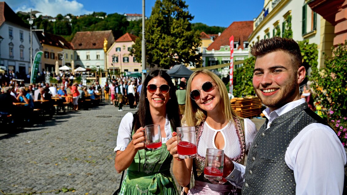 Drei Freunde in traditioneller Tracht halten Getränke in der Hand und lächeln. Im Hintergrund ist eine lebhafte Veranstaltung mit vielen Menschen und bunten Ständen zu sehen. | © Oststeiermark Tourismus