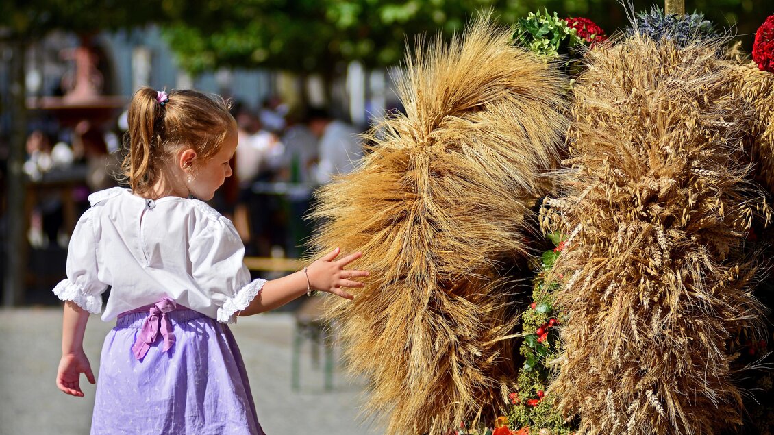 Ein kleines Mädchen mit Zöpfen berührt ein dekoratives Element aus Stroh. Im Hintergrund sind Menschen und Pflanzen zu sehen. | © Oststeiermark Tourismus