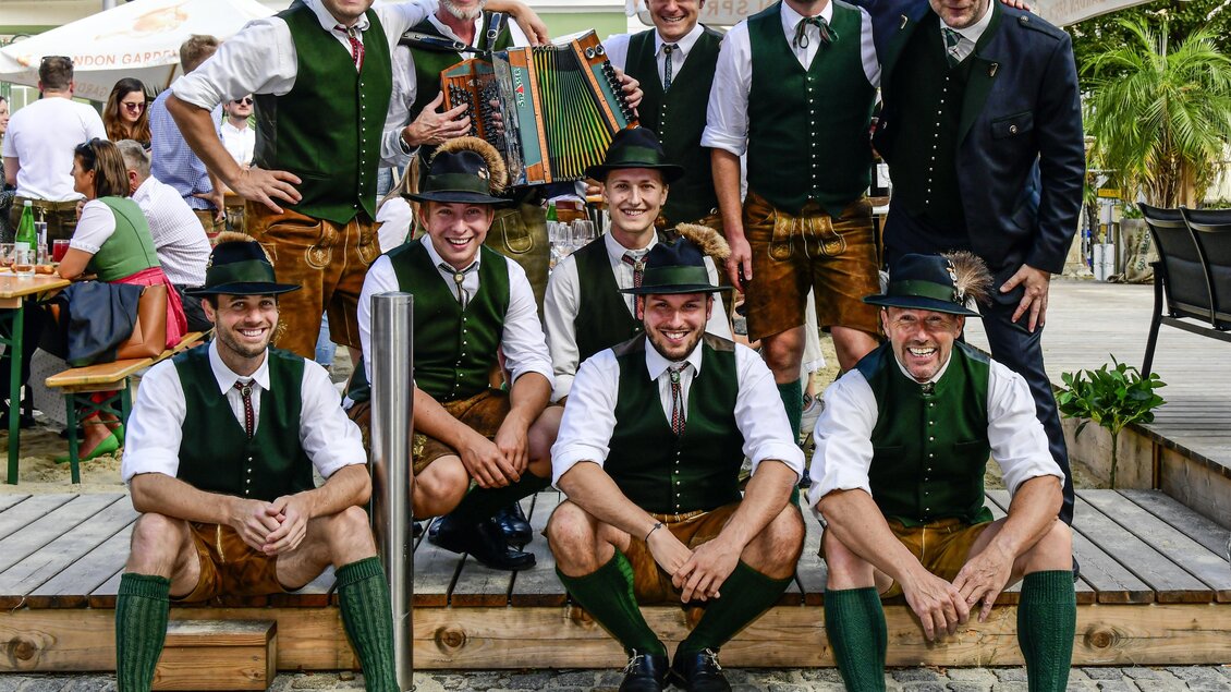 Eine Gruppe von Männern trägt traditionelle Trachten und steht in einer fröhlichen Atmosphäre. Sie posieren vor einem Restaurant mit hintergründigen Gästen und einem Holzdeck. | © Zugschwert