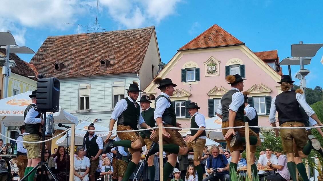 Eine Gruppe von Tänzern in traditioneller Tracht führt einen Volkstanz auf einer Bühne vor bunt gestalteten Häusern auf. Im Hintergrund sitzen viele Zuschauer und genießen die Aufführung. | © Stadtgemeinde Hartberg