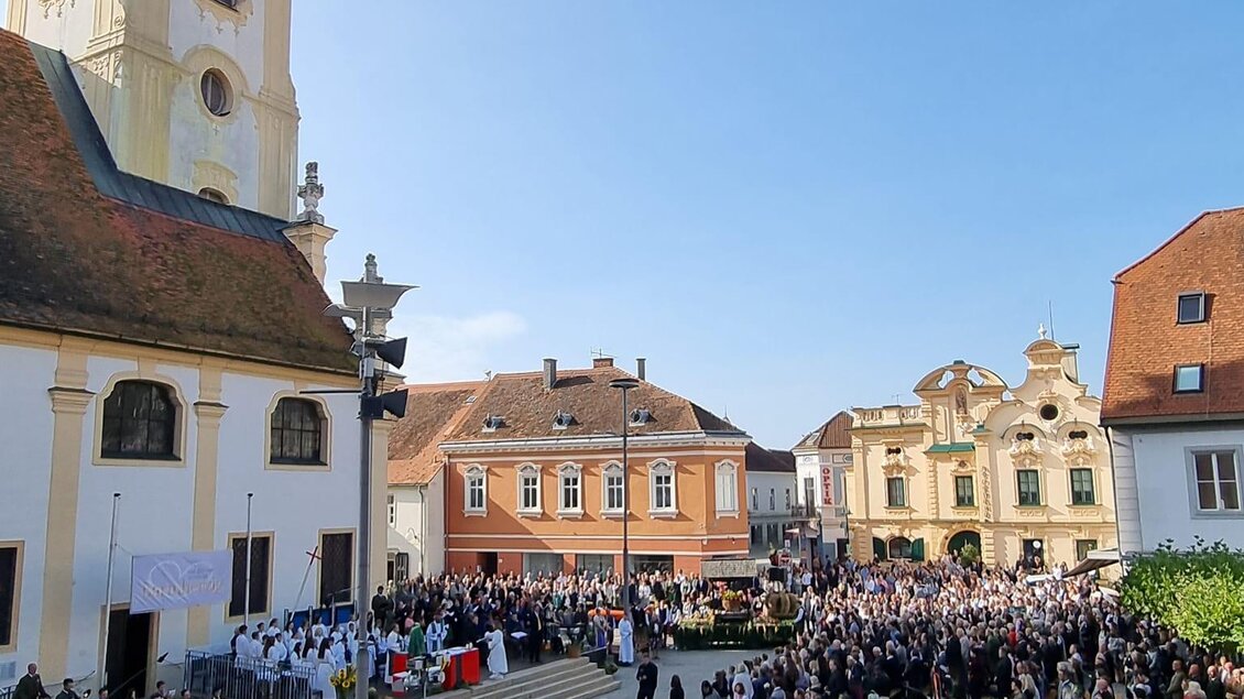 Eine große Menschenmenge versammelt sich auf einem Platz vor einer Kirche. Im Hintergrund sind historische Gebäude und ein klarer blauer Himmel zu sehen. | © Stadtgemeinde Hartberg