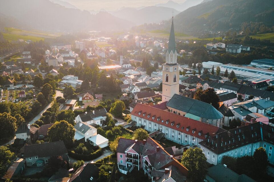 Eine malerische Stadt in den Bergen mit einer Kirche und vielen bunten Häusern. Die Sonne geht hinter den Hügeln unter und taucht die Landschaft in warmes Licht.