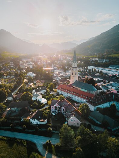 Eine malerische Stadt in den Bergen mit einer Kirche und vielen bunten Häusern. Die Sonne geht hinter den Hügeln unter und taucht die Landschaft in warmes Licht.