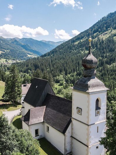 Eine malerische Kirche in den Bergen, umgeben von grünen Wiesen und dichten Wäldern. Der Himmel ist klar und die Landschaft strahlt Ruhe aus.