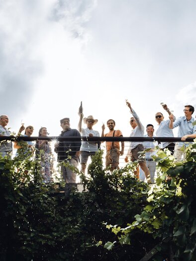 A group of people is standing on a terrace and waving happily. Behind them, there are green plants and a cloudy sky. | © Katrin Meitz