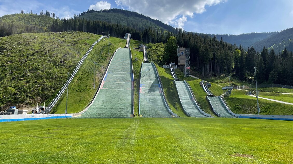 Eine Sprungschanze in den Bergen mit grünem Gras davor. Im Hintergrund sind Wälder und ein blauer Himmel zu sehen. | © NAZ Eisenerz
