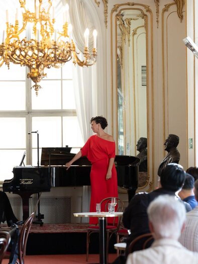 An elegant performance in a stylish room with chandeliers and large windows. Two singers stand at the piano and entertain an audience. | © Oper Graz