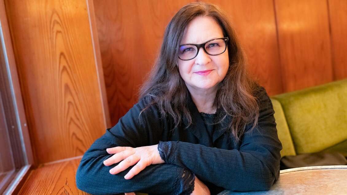 Eine freundliche Frau, die Autorin Bettina Balàka, schaut freundlich in die Kamera. Sie hat lange brünette Haare, trägt eine Brille und hat die Arme verschränkt auf einen Tisch gelegt.  
 | © Die Wasnerin, www.diewasnerin.at
