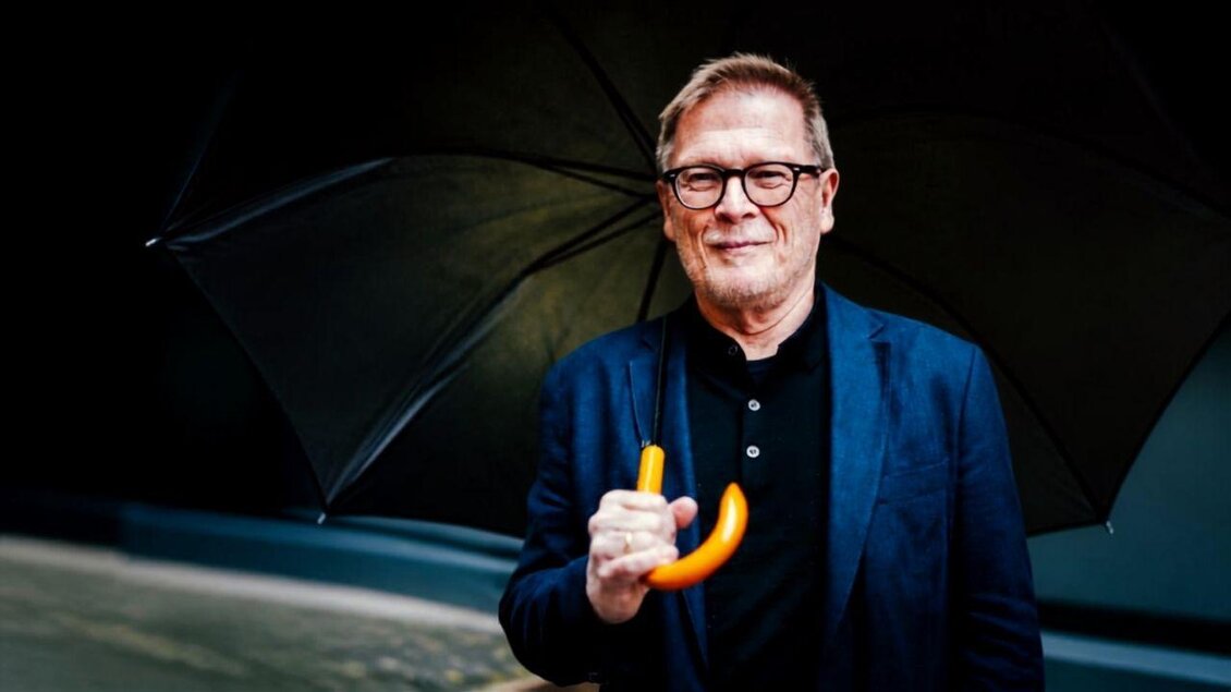 <p>Ein Bild des Autors und Vortragenden Paulus Hochgatterer, welcher im Hotel Die Wasnerin aus seinem Roman vorliest. Er steht draußen im Freien an einem Regentag und hält einen Regenschirm in der Hand und lacht. | © Die Wasnerin